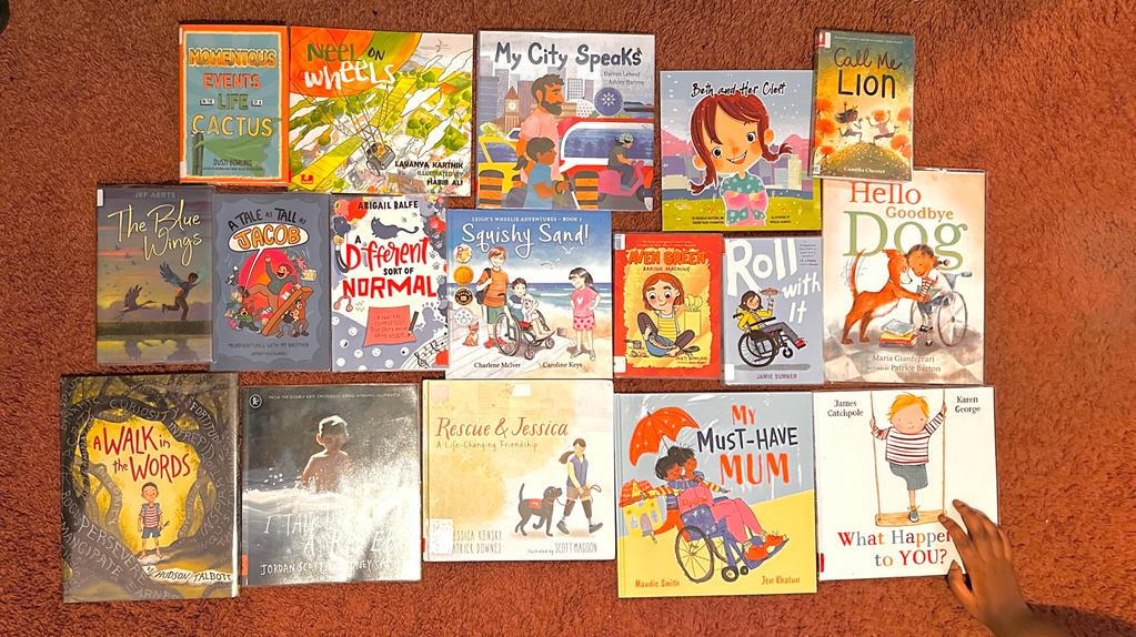 This picture is 16 different Disability children's books spread out across the floor in my room. My hand is in the corner holding one of them.